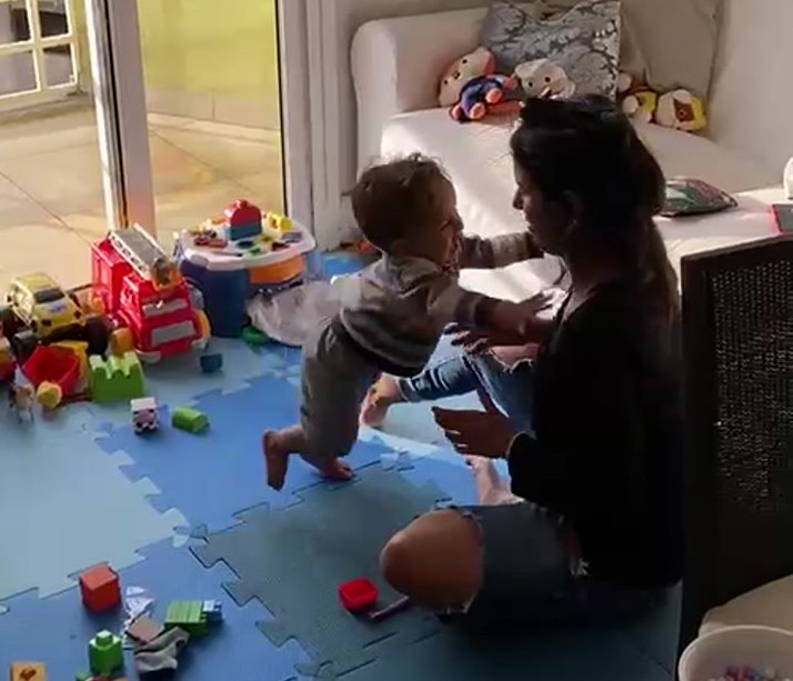 Menino dando os primeiros passos em direção à mãe, na sala de casa.